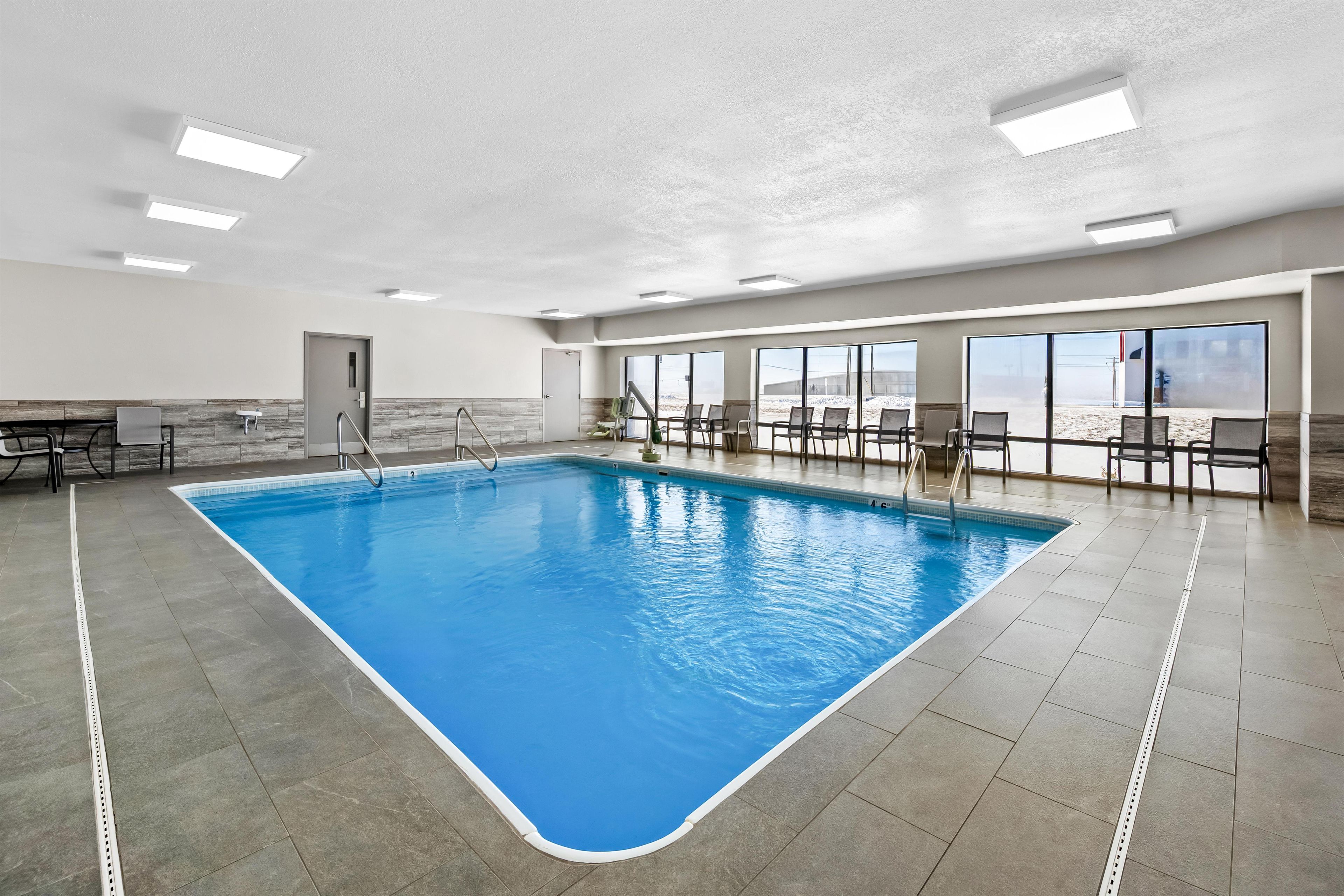 An indoor swimming pool with clear blue water, surrounded by a tiled floor and walls, with a seating area and large windows.