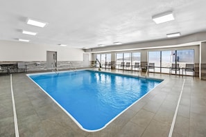An indoor swimming pool with clear blue water, surrounded by a tiled floor and walls, with a seating area and large windows.