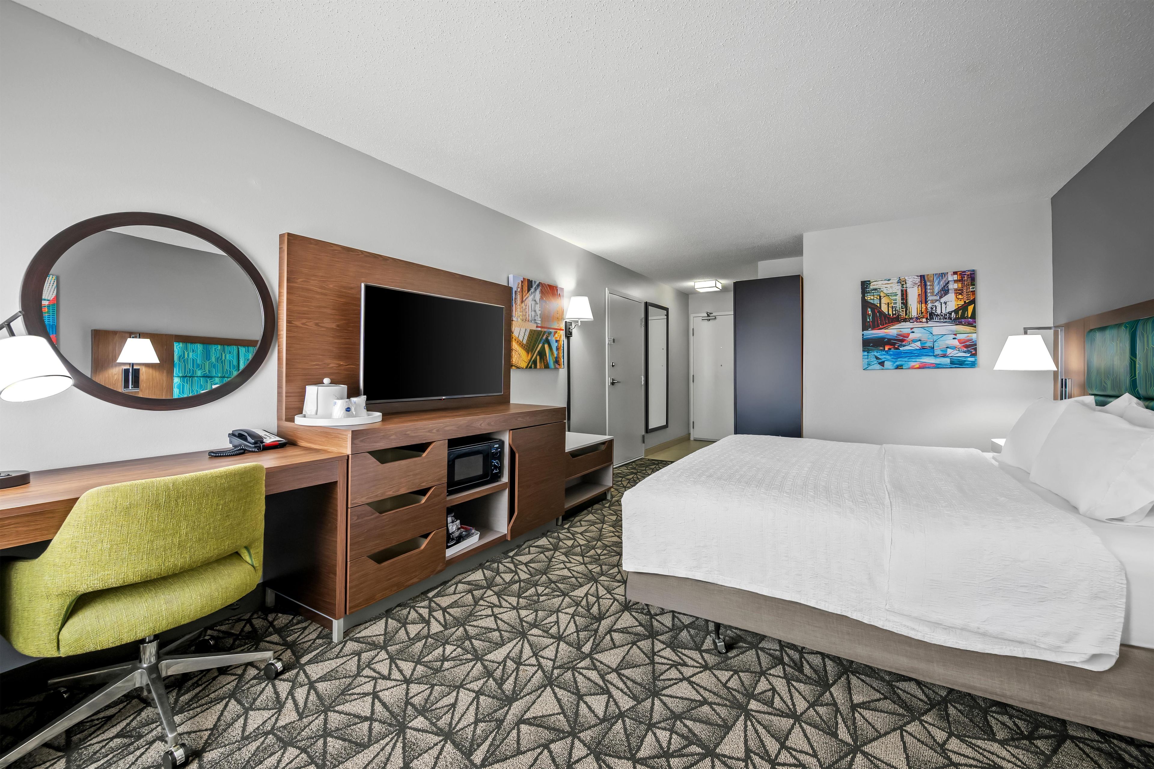 Ein Hotelzimmer mit einem Bett, einem Schreibtisch, einem Flachbildfernseher, einem Stuhl, einem Spiegel und Kunstwerken an den Wänden.