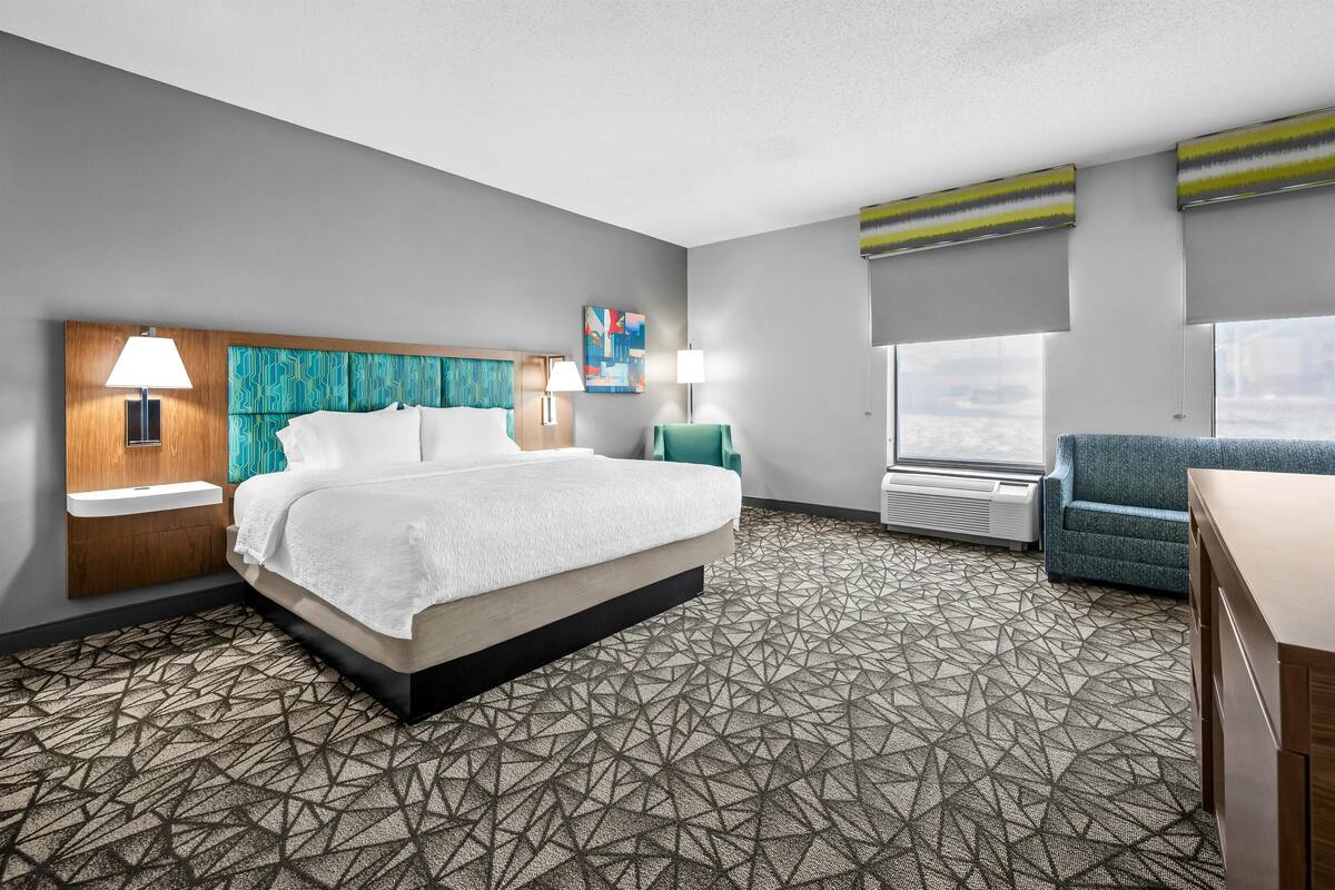 A hotel room with a bed, two bedside tables with lamps, a desk, a chair, a window, and a painting on the wall.