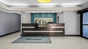 A reception area with a 'WELCOME' mat, a reception desk, and a view of industrial buildings through the window. - Hampton Inn Bloomington West (Bloomington)