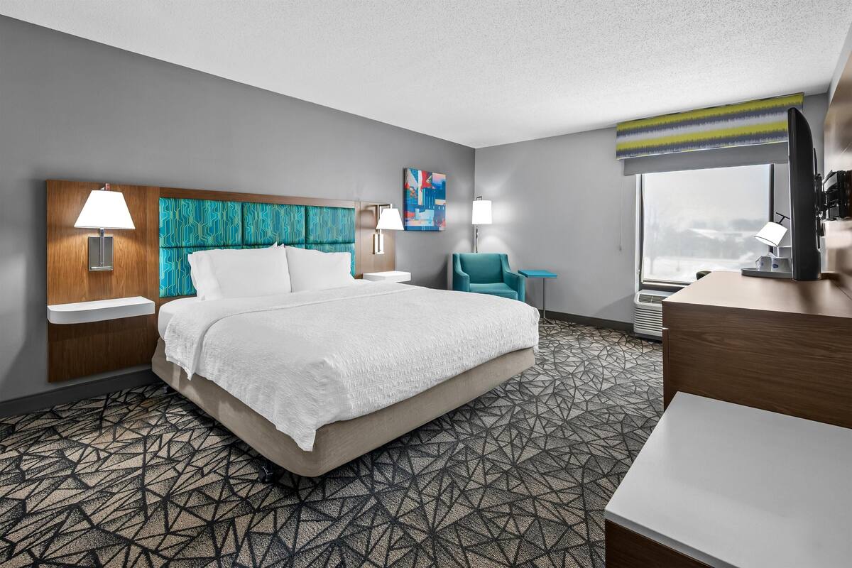 A hotel room with a large bed, two bedside tables with lamps, a desk, a chair, and a large window with a view.