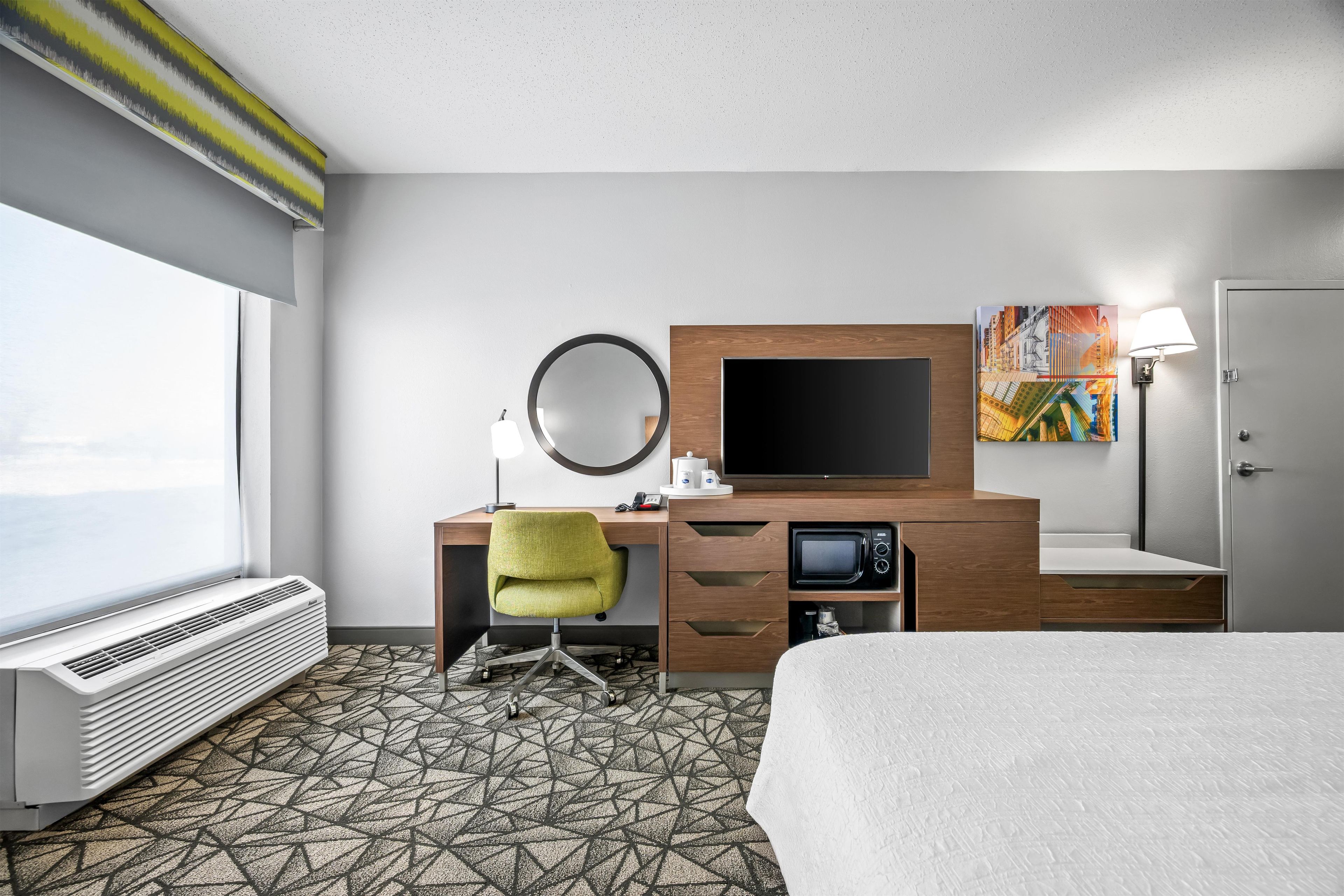 A hotel room with a bed, a desk with a computer, a microwave, a TV, and a window with a view.