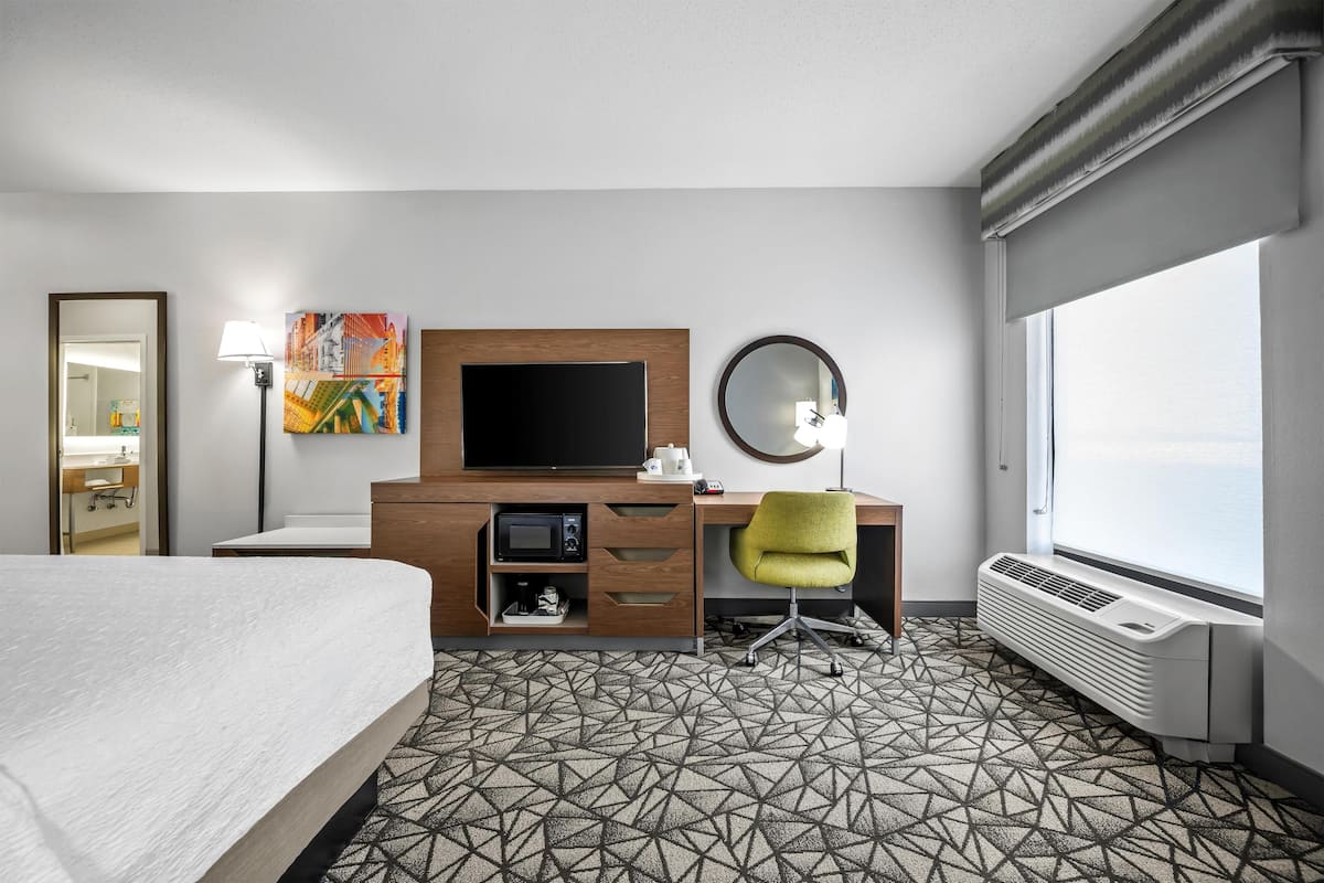 A hotel room with a bed, a desk with a flat-screen TV, a microwave, a chair, a large window, and a patterned floor.