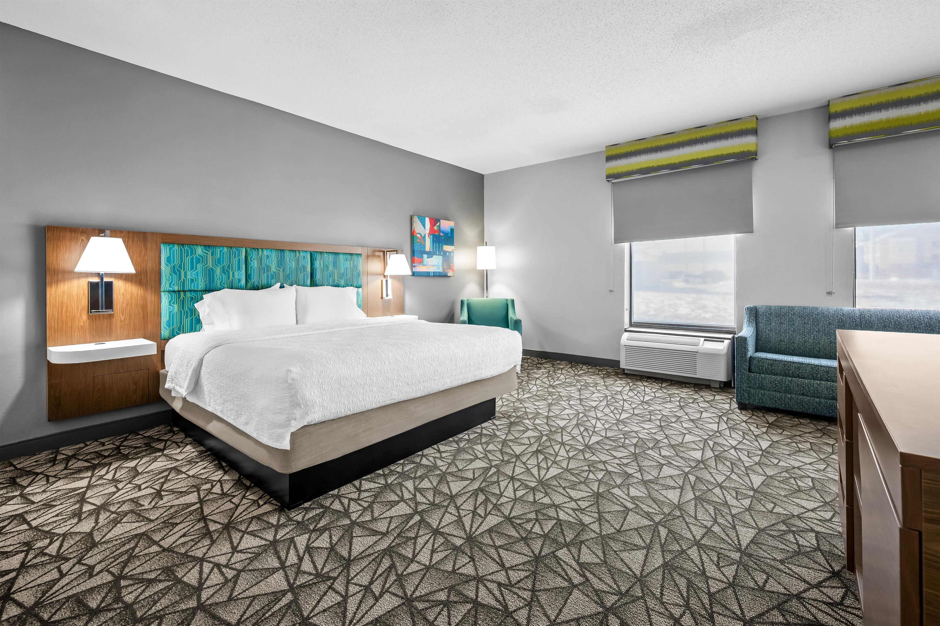A hotel room with a bed, two bedside lamps, a painting, a window with a view, and a seating area with a chair.
