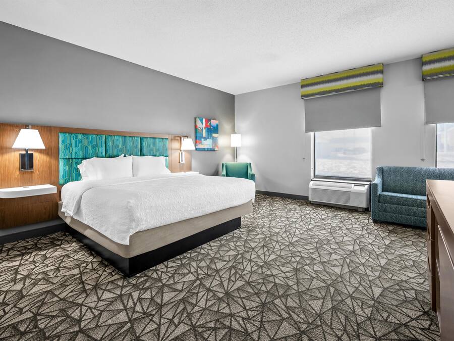 A hotel room with a bed, two bedside lamps, a painting, a window with a view, and a seating area with a chair.