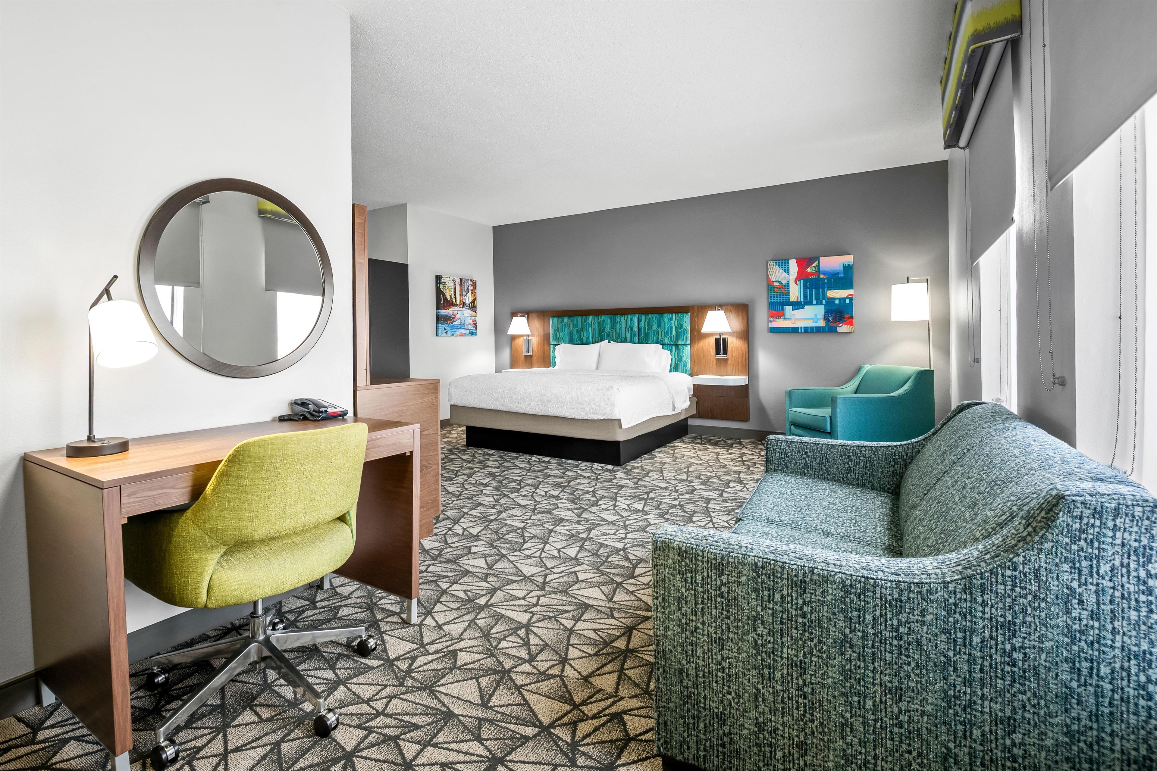 Habitación de hotel con cama, escritorio con silla, sofá, espejo y obras de arte en las paredes.
