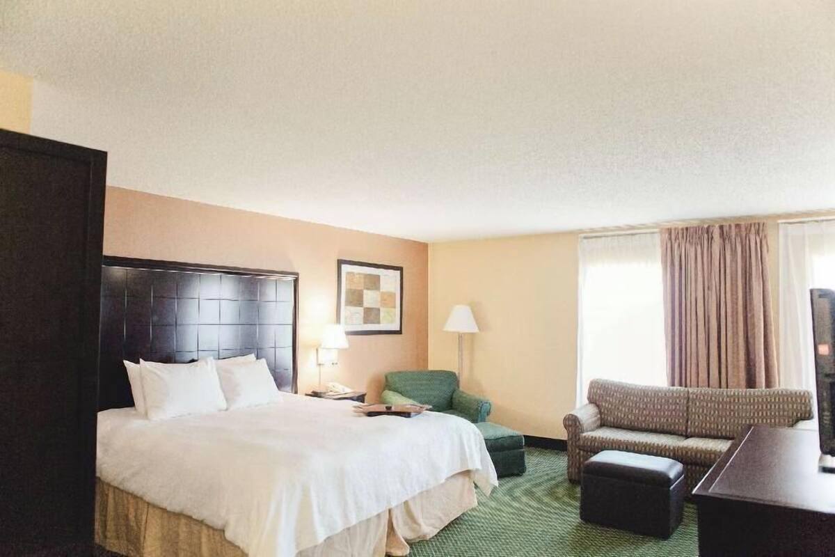 A hotel room with a large bed, a green armchair, a sofa, a desk, and a television.