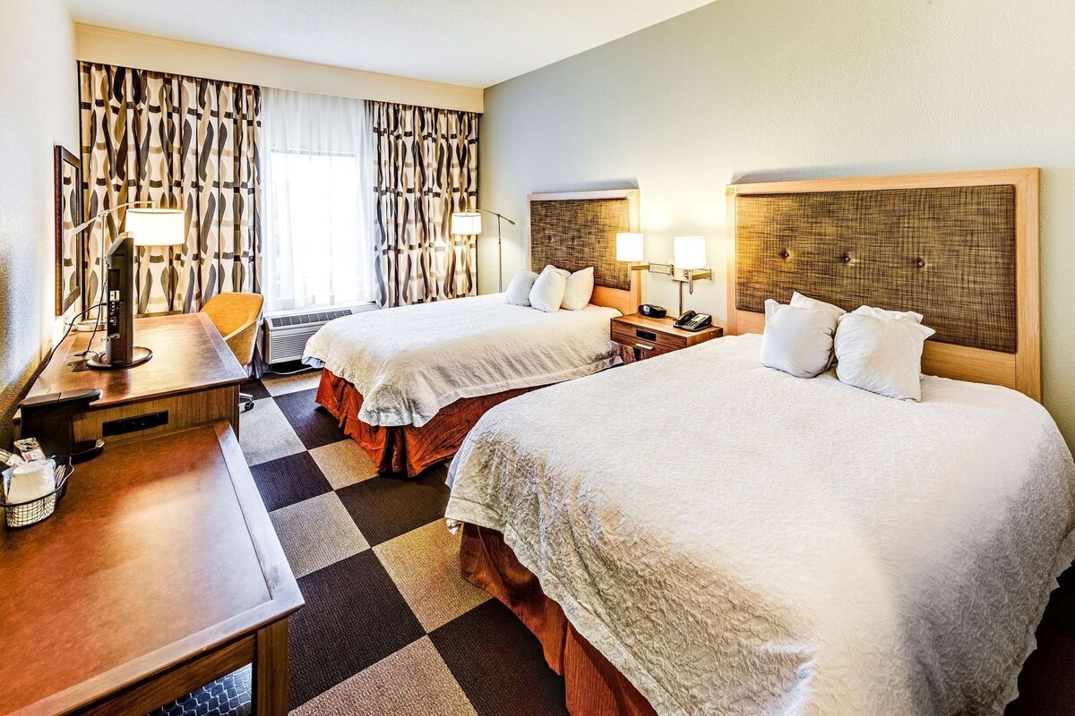 A hotel room with two beds, a desk, a chair, a lamp, and a window with curtains.