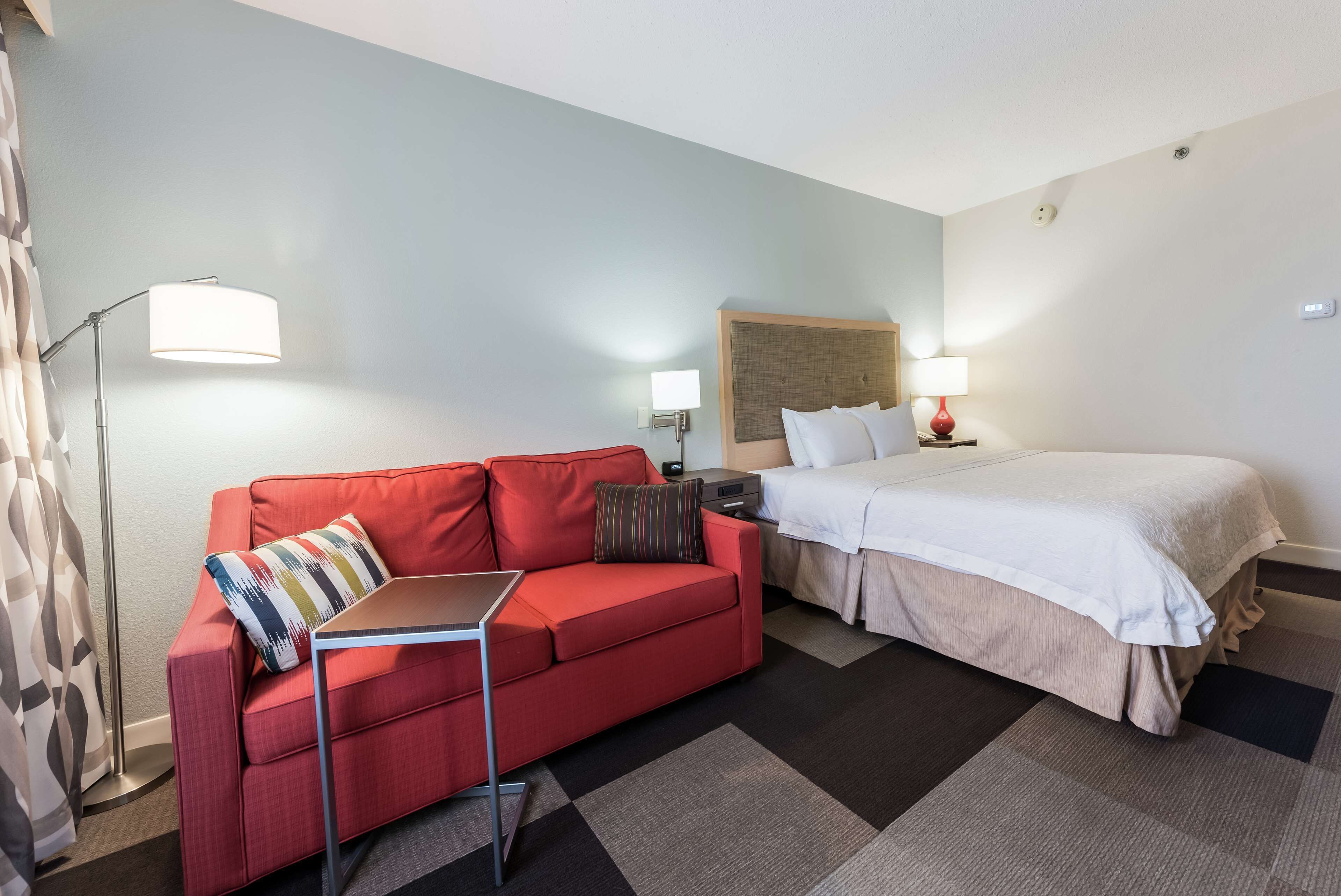 A hotel room with a red sofa, a bed with white bedding, a small table, and a floor lamp.
