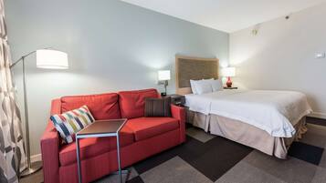 A hotel room with a red sofa, a bed with white bedding, a small table, and a floor lamp.