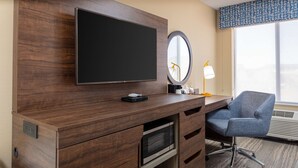 A hotel room with a wooden desk, a flat-screen TV, a microwave, a chair, a mirror, and a window with a view.