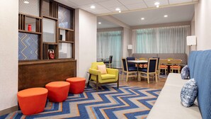 A hotel lobby with a blue and orange chevron-patterned carpet, a yellow armchair, red ottomans, a blue sofa, and a dining area with wooden chairs and a table.