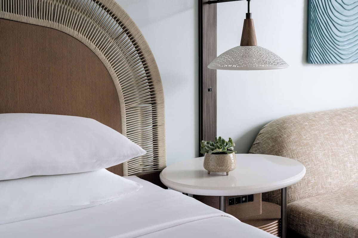 A neatly arranged hotel room with a bed, a round table with a potted plant, a hanging lamp, and a wall-mounted artwork.