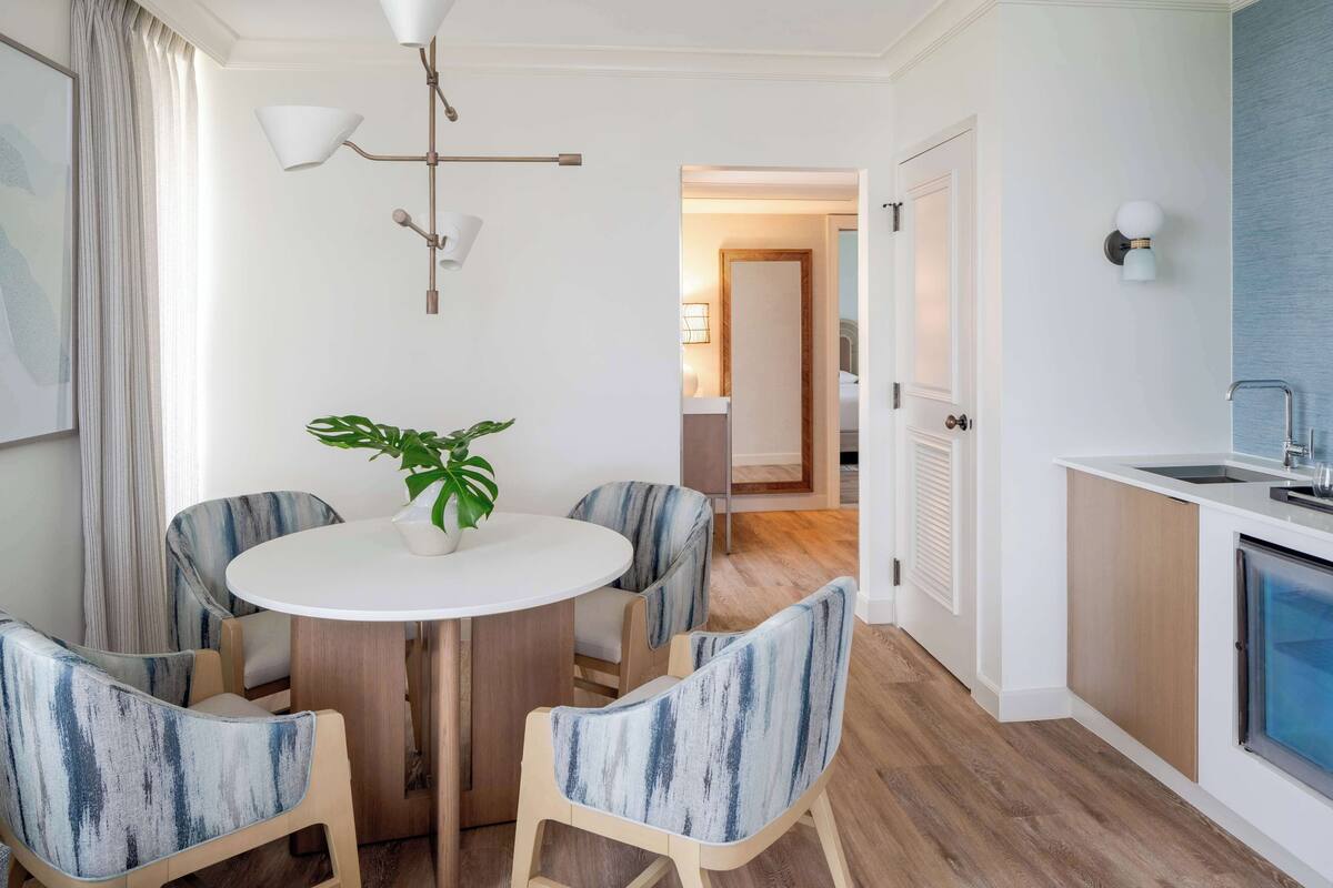 A modern kitchen with a white countertop, a sink, and a built-in oven. There is a dining area with a round table and four chairs, each with a blue and white patterned cushion. A potted plant is on the table.