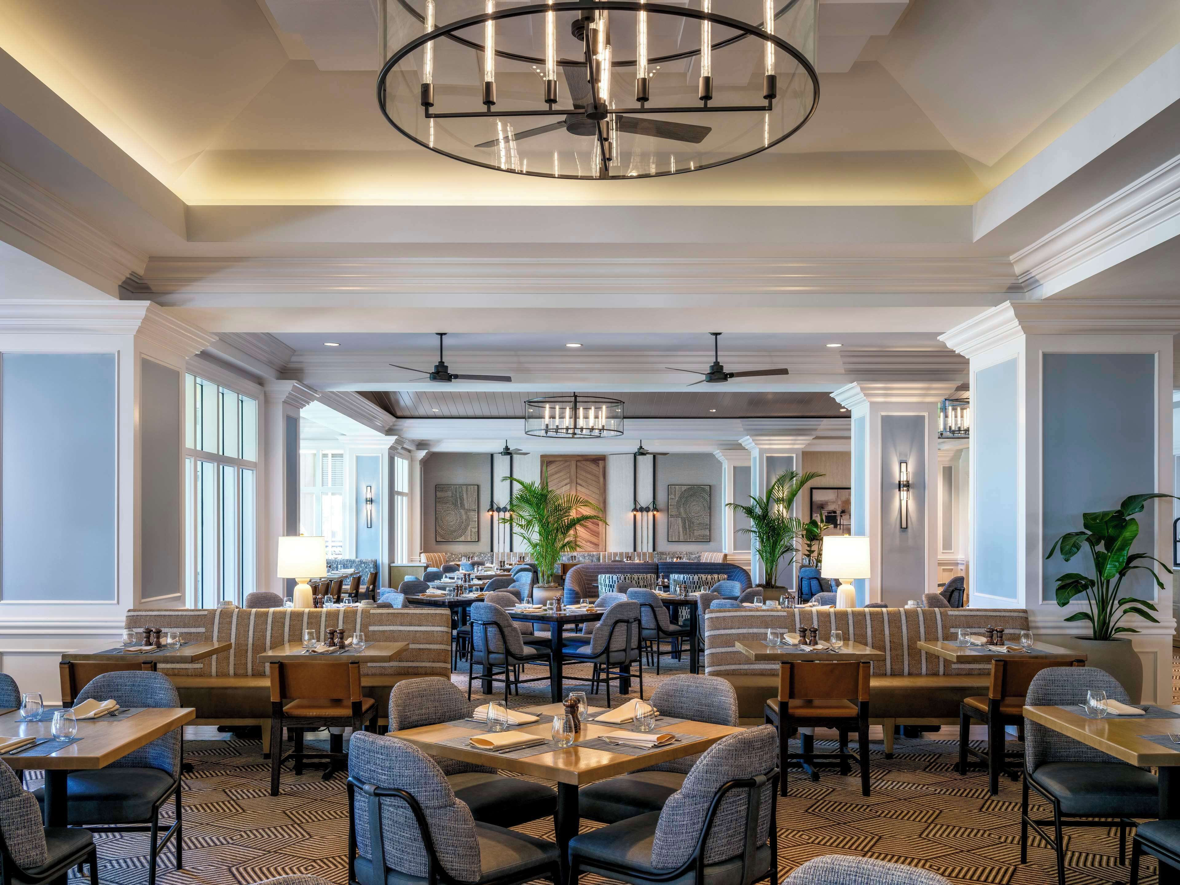A spacious dining area with multiple tables and chairs, a central chandelier, and ceiling fans.