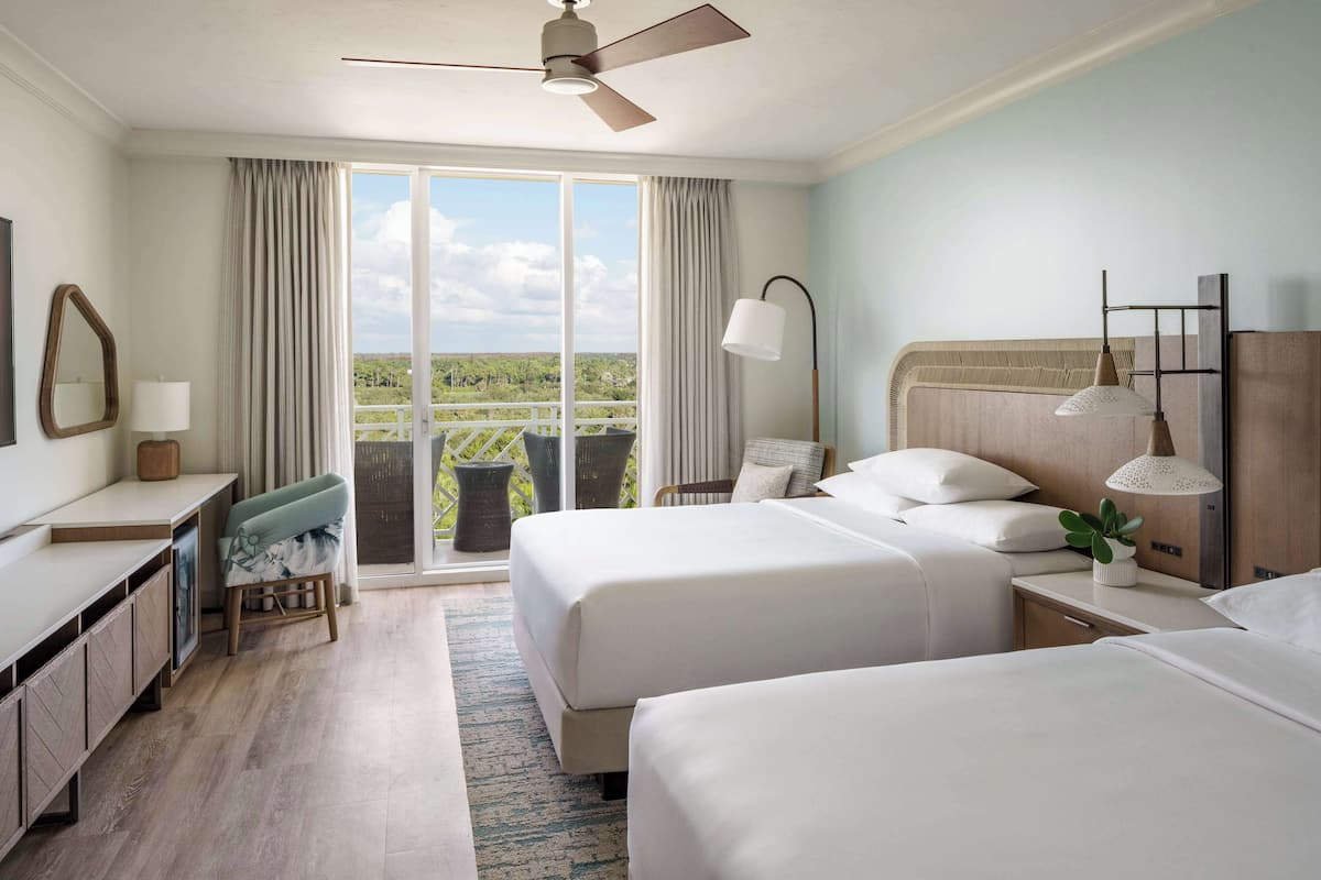 A hotel room with two beds, a desk, a chair, a balcony with a view, and a ceiling fan.