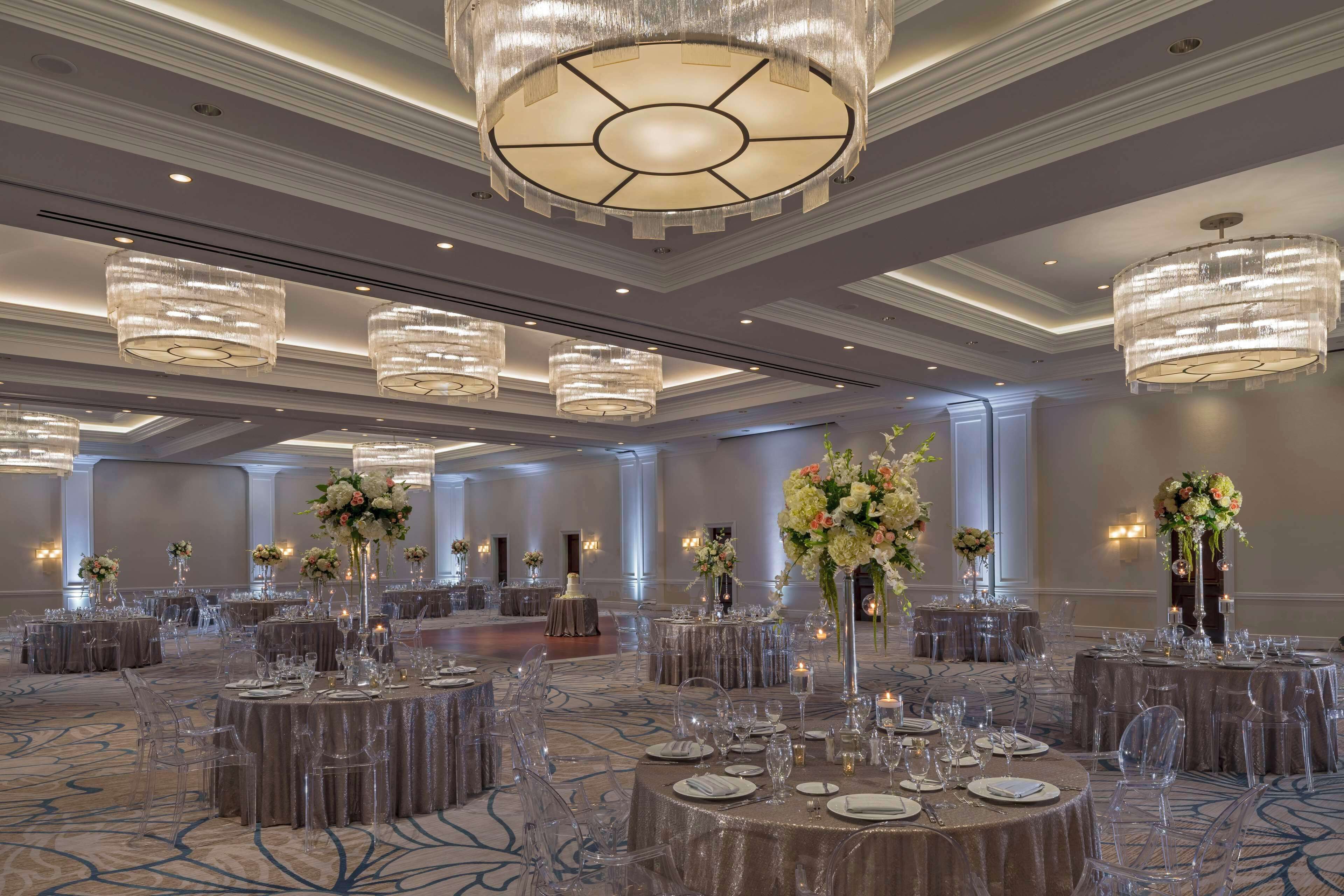 A spacious banquet hall with round tables set for events, featuring clear chairs, elegant table settings, and large chandeliers.