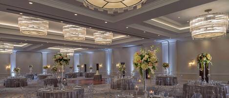 Un amplio salón de banquetes con mesas redondas dispuestas para eventos, sillas traslúcidas, elegantes centros de mesa y grandes candelabros.