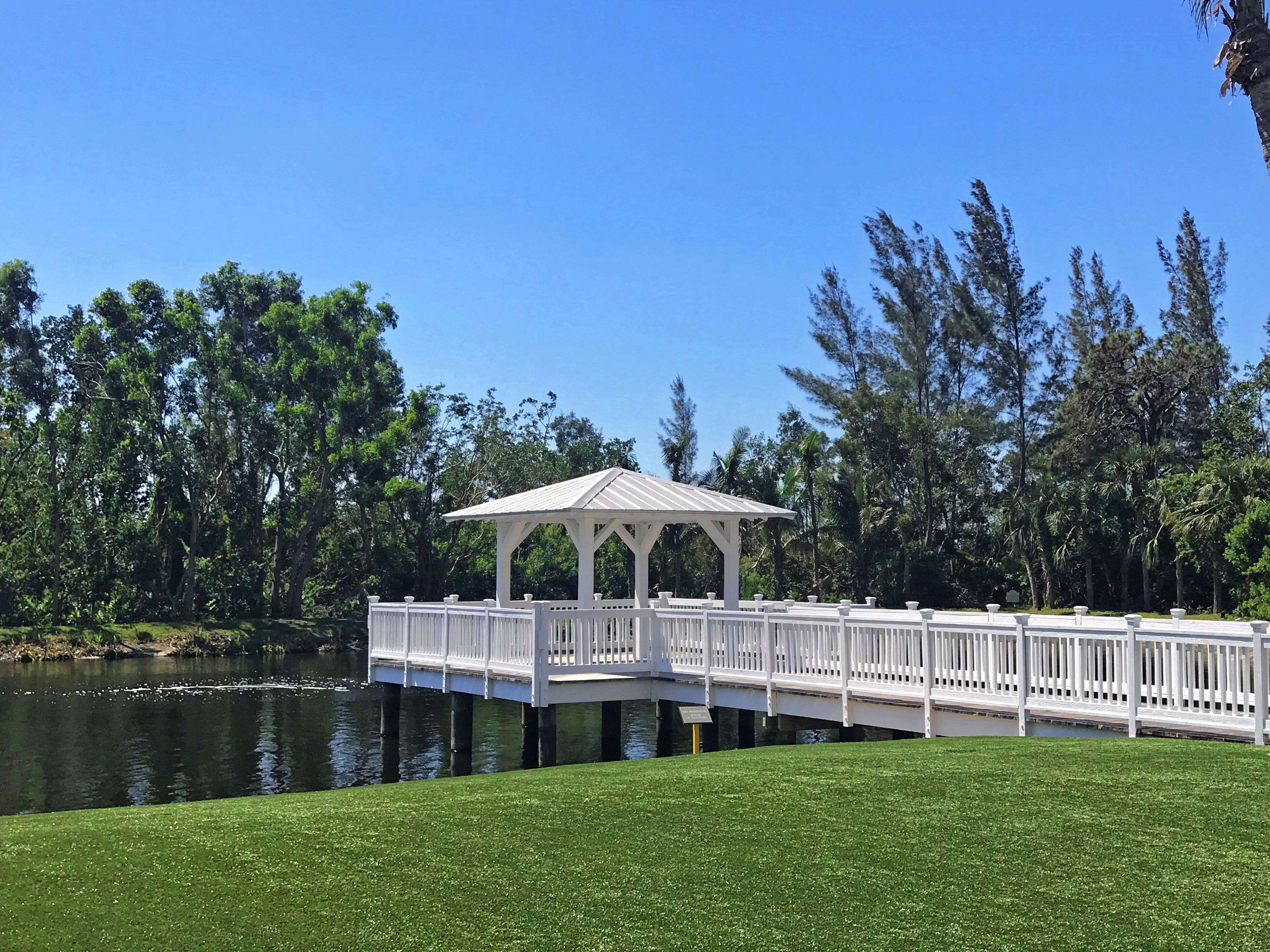 Een witte pergola met dak op een houten terras boven water, omgeven door weelderige begroeiing.
