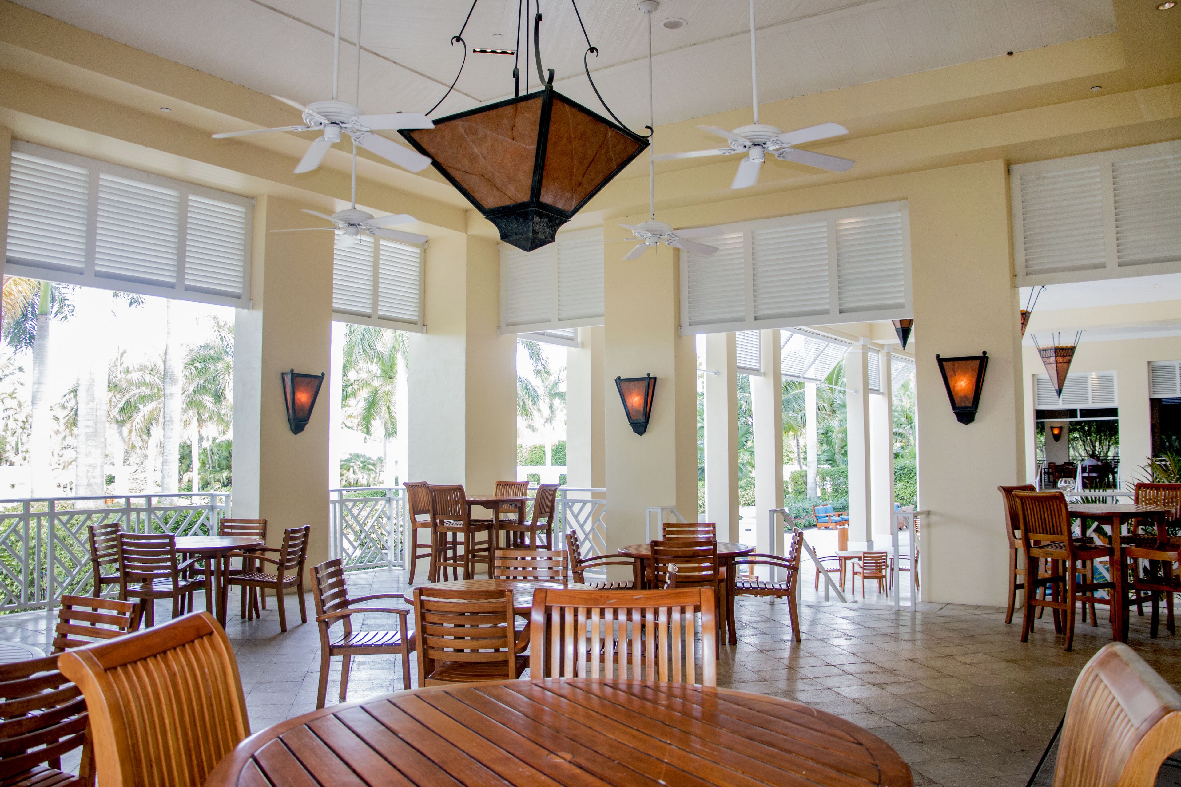 A spacious indoor area with multiple wooden tables and chairs, ceiling fans, and large windows allowing natural light.
