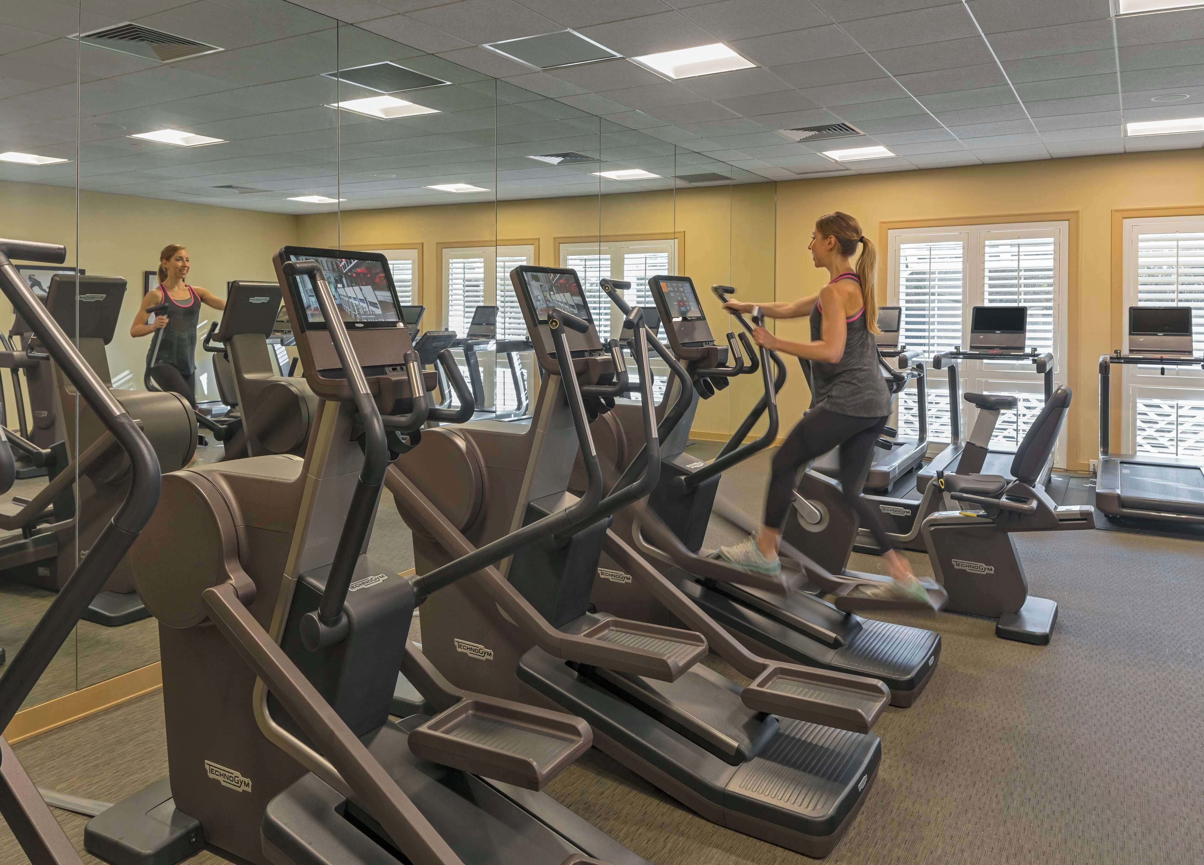 A gym with multiple treadmills and exercise machines, large windows with blinds, and a mirrored wall.