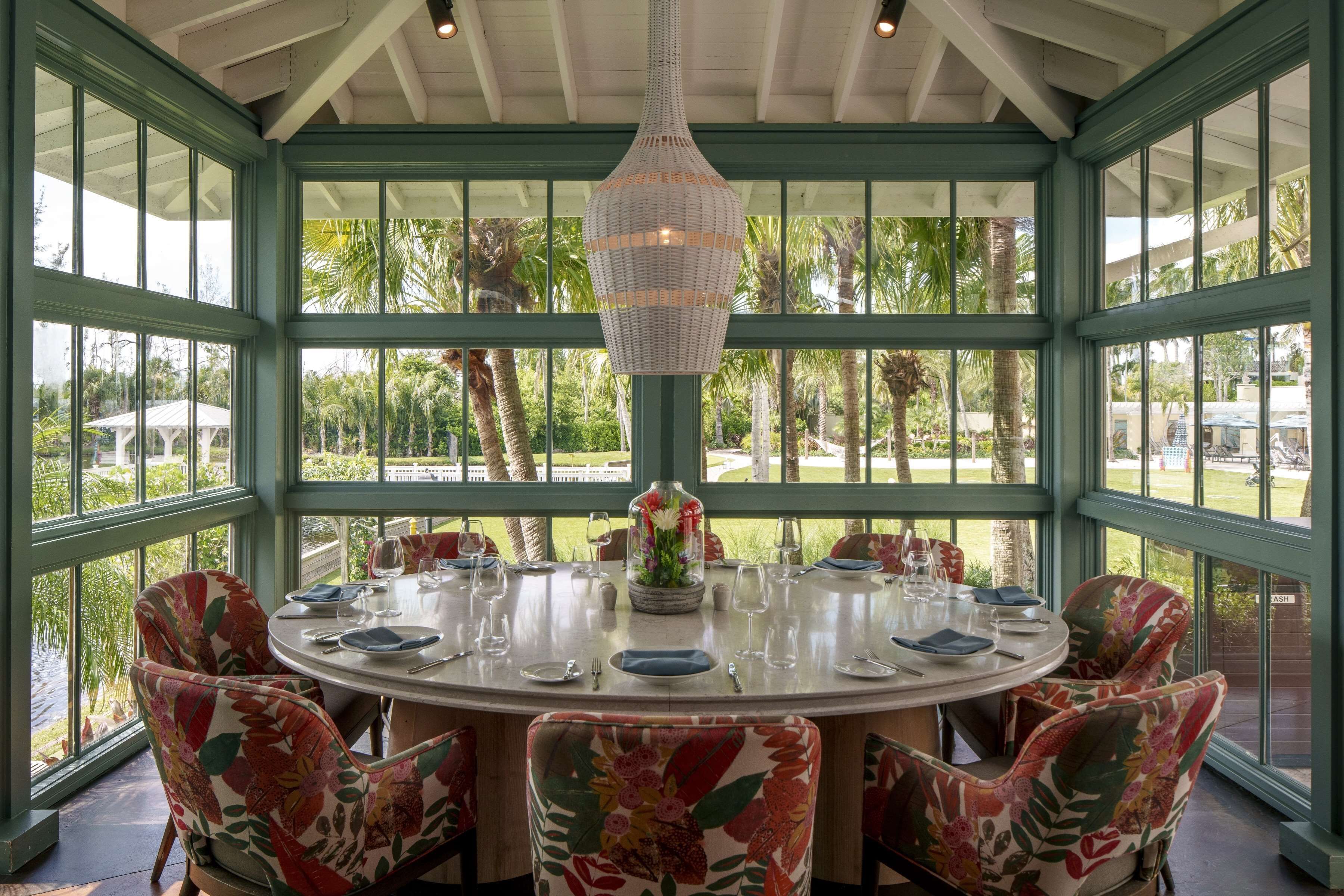 A round dining table with floral chairs, a hanging light fixture, and a view of palm trees through large windows.