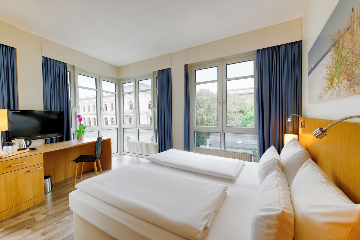 Una habitación de hotel con cama, escritorio, silla, televisor y ventanas amplias.