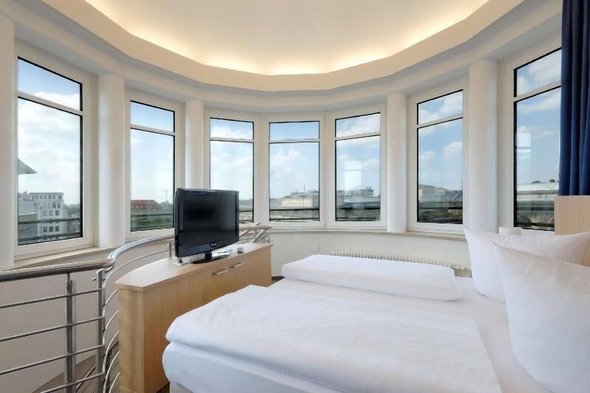 Une chambre d’hôtel avec un plafond incurvé, de grandes fenêtres, un lit, une télévision et une commode en bois.