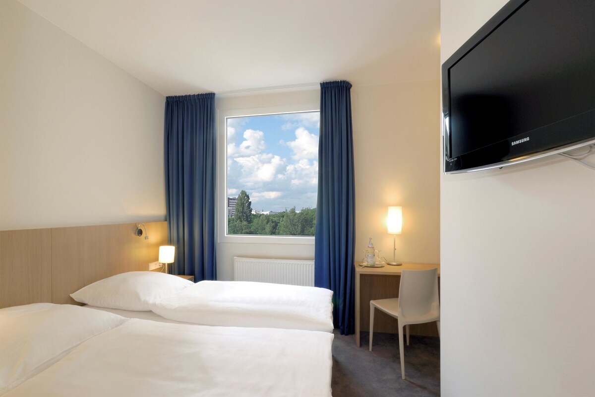 Habitación de hotel con cama, escritorio, silla, televisor y ventana con cortinas.