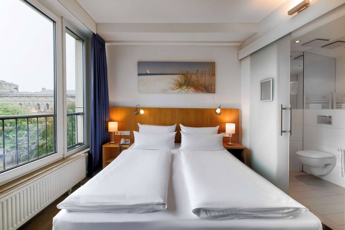 Une chambre d’hôtel avec deux lits, une vue sur les bâtiments environnants, un tableau au mur et une salle de bain avec des toilettes et une douche.