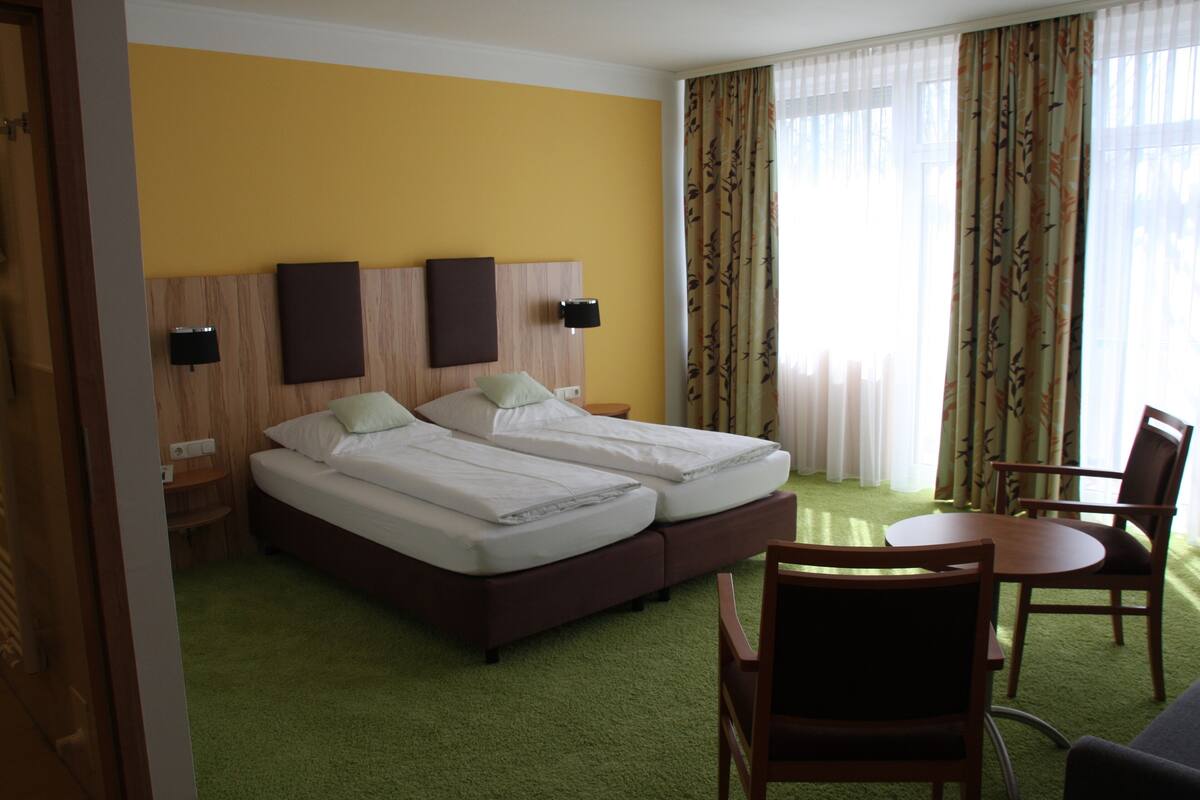 Et hotelværelse med to senge, grønt tæppe, trægavl, et lille bord og en stol.