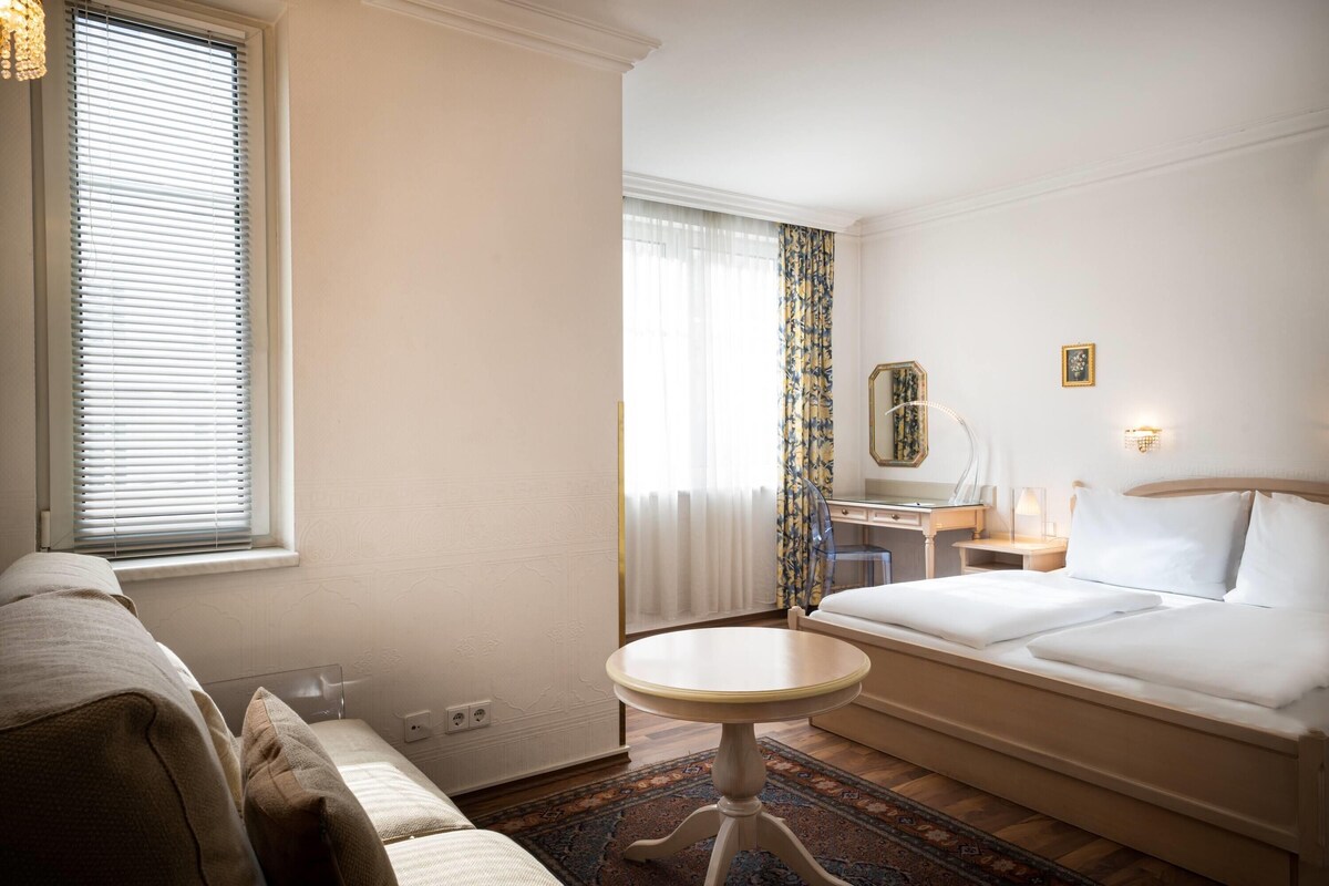 A hotel room with a bed, a desk, a chair, a small round table, and a window with blinds.