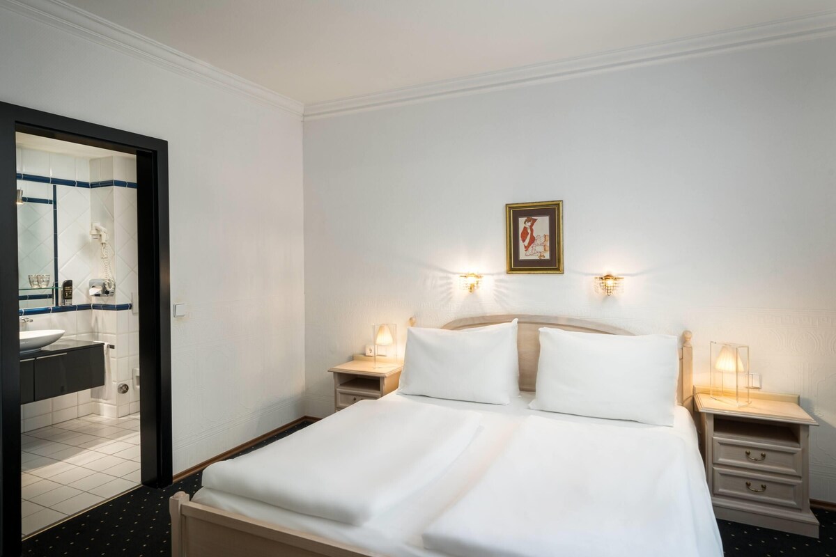 A hotel room with a bed, bedside tables, a nightstand, a mirror, and a framed picture on the wall.