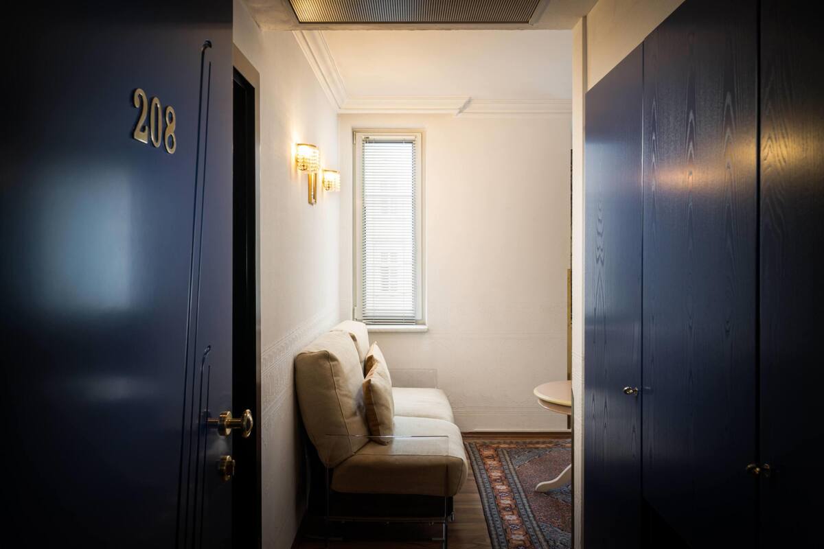A hallway with a door labeled '208,' a beige sofa with cushions, a small round table, and a window with blinds.