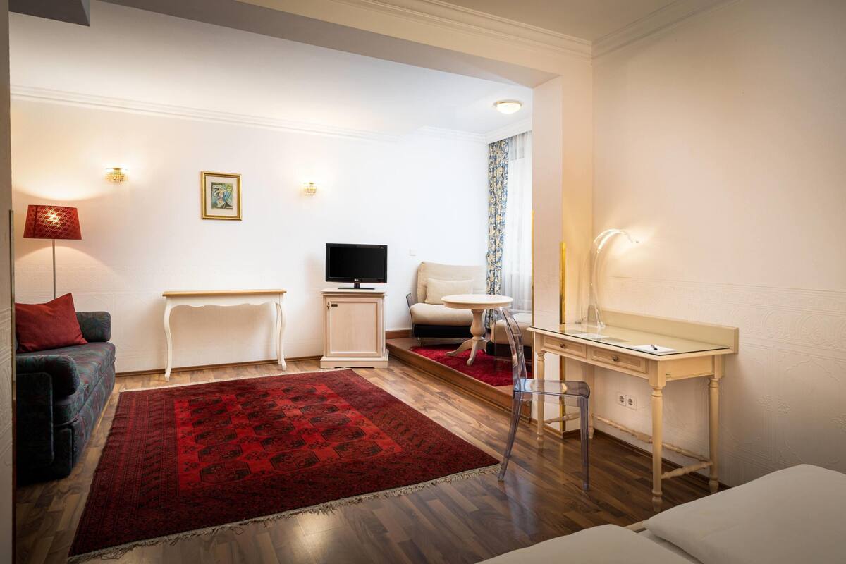 A hotel room with a sofa, a TV, a desk, a chair, a red rug, and a bed.