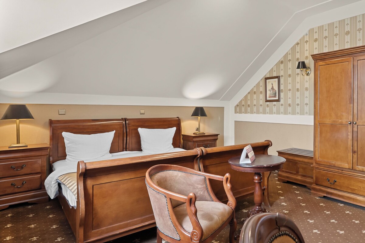 A traditional bedroom with a wooden bed, two bedside tables with lamps, a wooden chair, a small round table, and a wardrobe.