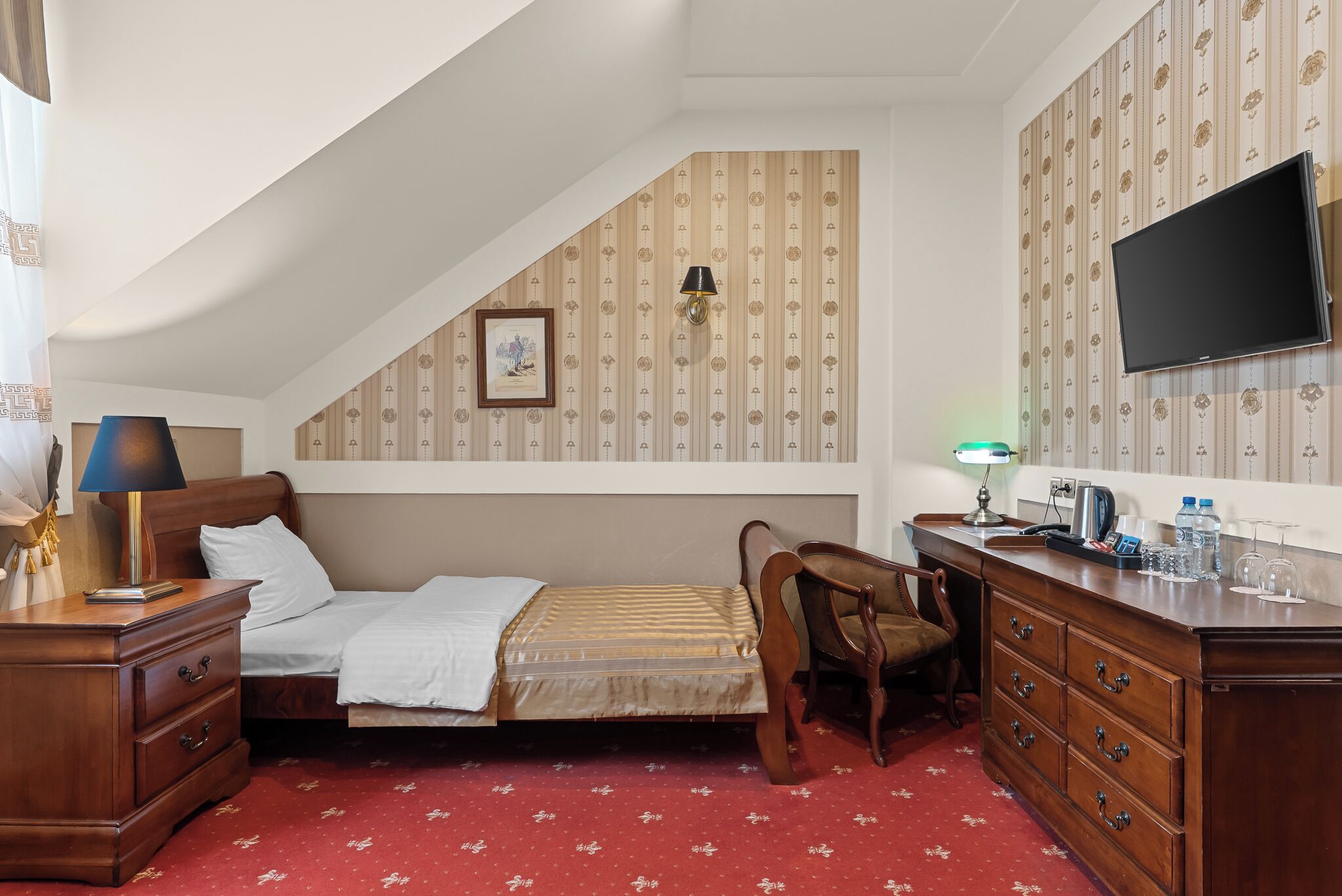 Habitación de hotel pequeña con una cama individual, un escritorio con silla, una cómoda, un televisor y una pared con diseño.