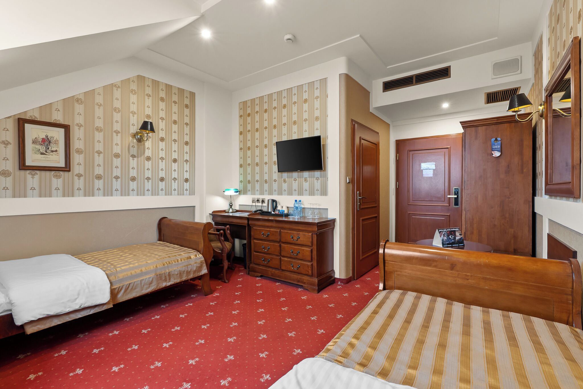 A hotel room with two single beds, a desk with a chair, a television, a wardrobe, and a red carpet.