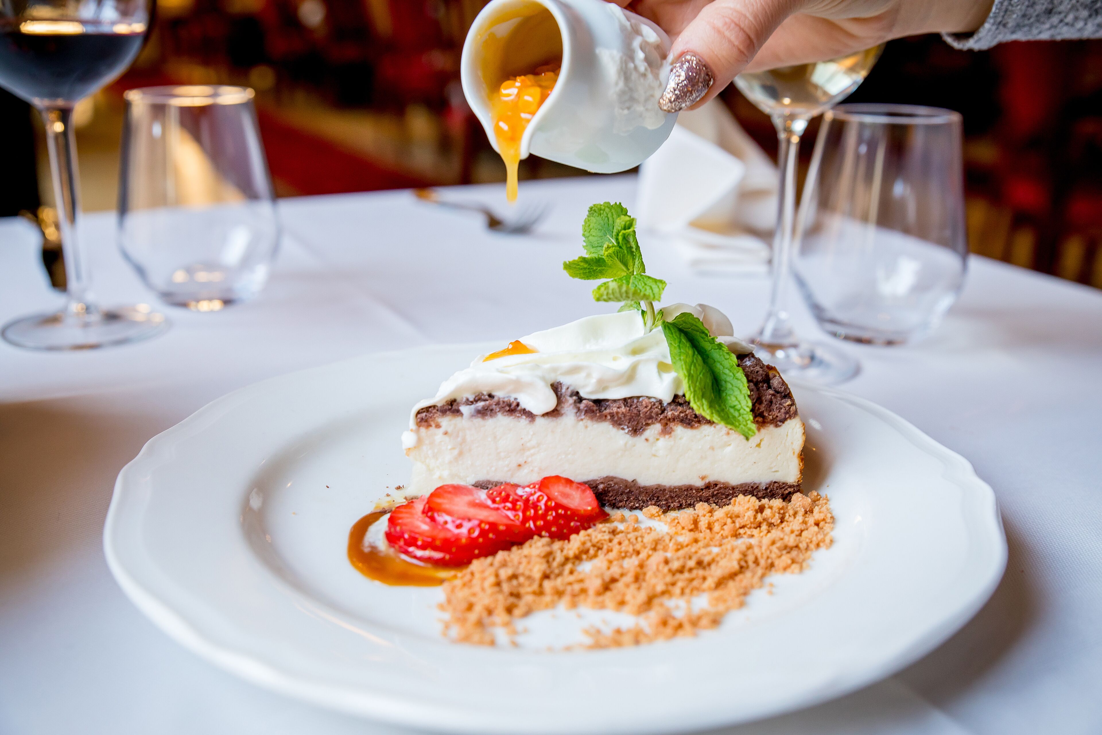 Un dessert servi dans une assiette blanche, décoré d’une feuille de menthe, de fraises et d’une garniture croustillante, et sur lequel une main verse de la sauce.