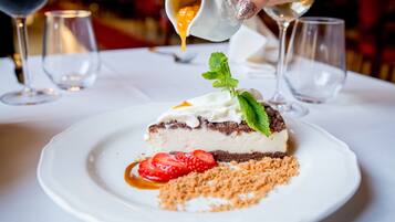 Dessert op een wit bord, gegarneerd met een muntblaadje, aardbeien en een kruimelige topping, terwijl er saus over gegoten wordt.