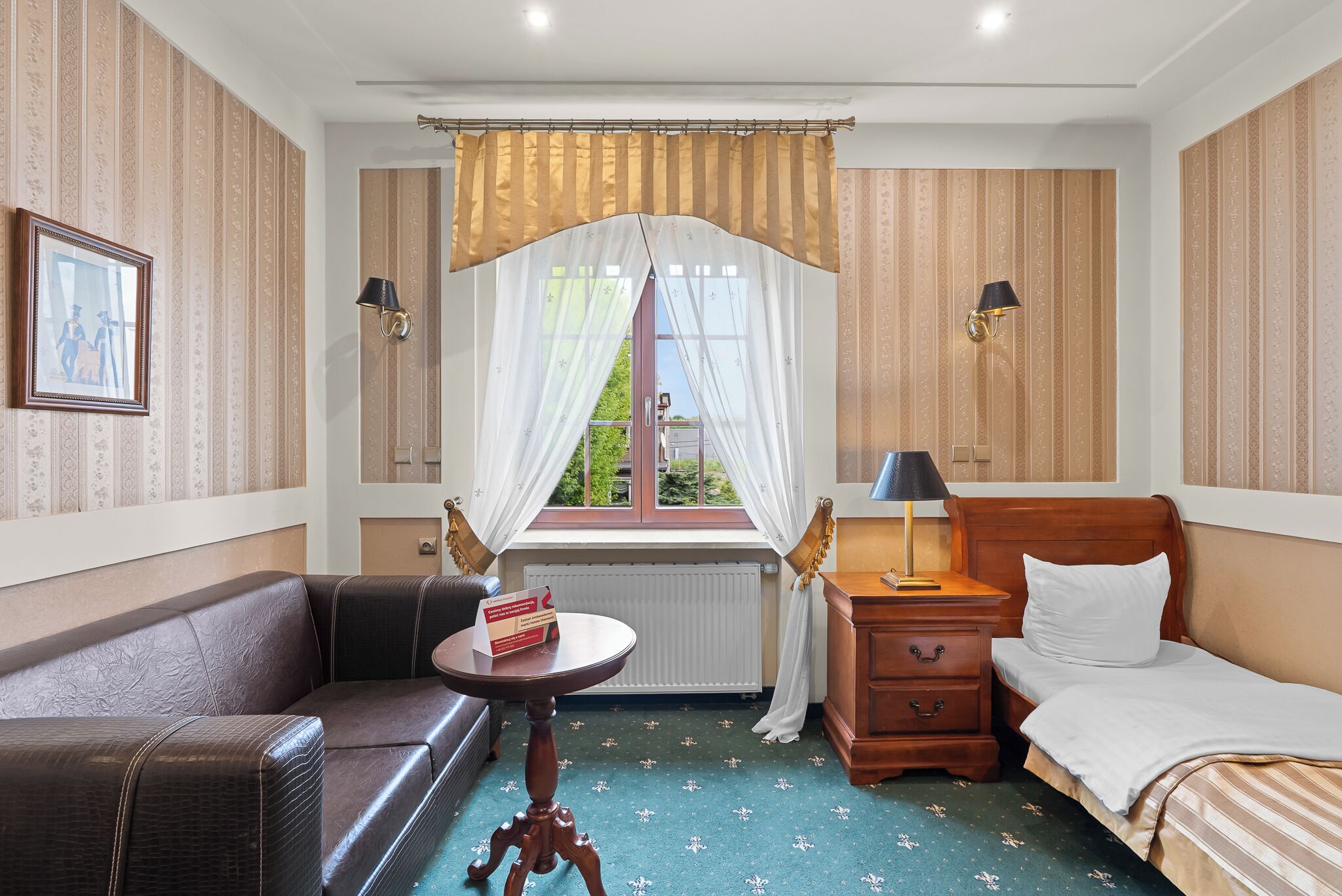 A hotel room with a bed, a small table, a nightstand, a sofa, and a window with curtains.