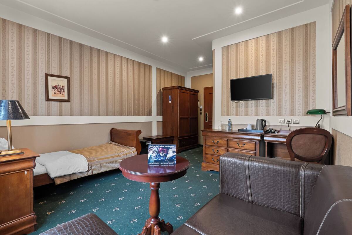 A hotel room with a bed, a small table, a desk with a chair, a wardrobe, and a television on the wall.
