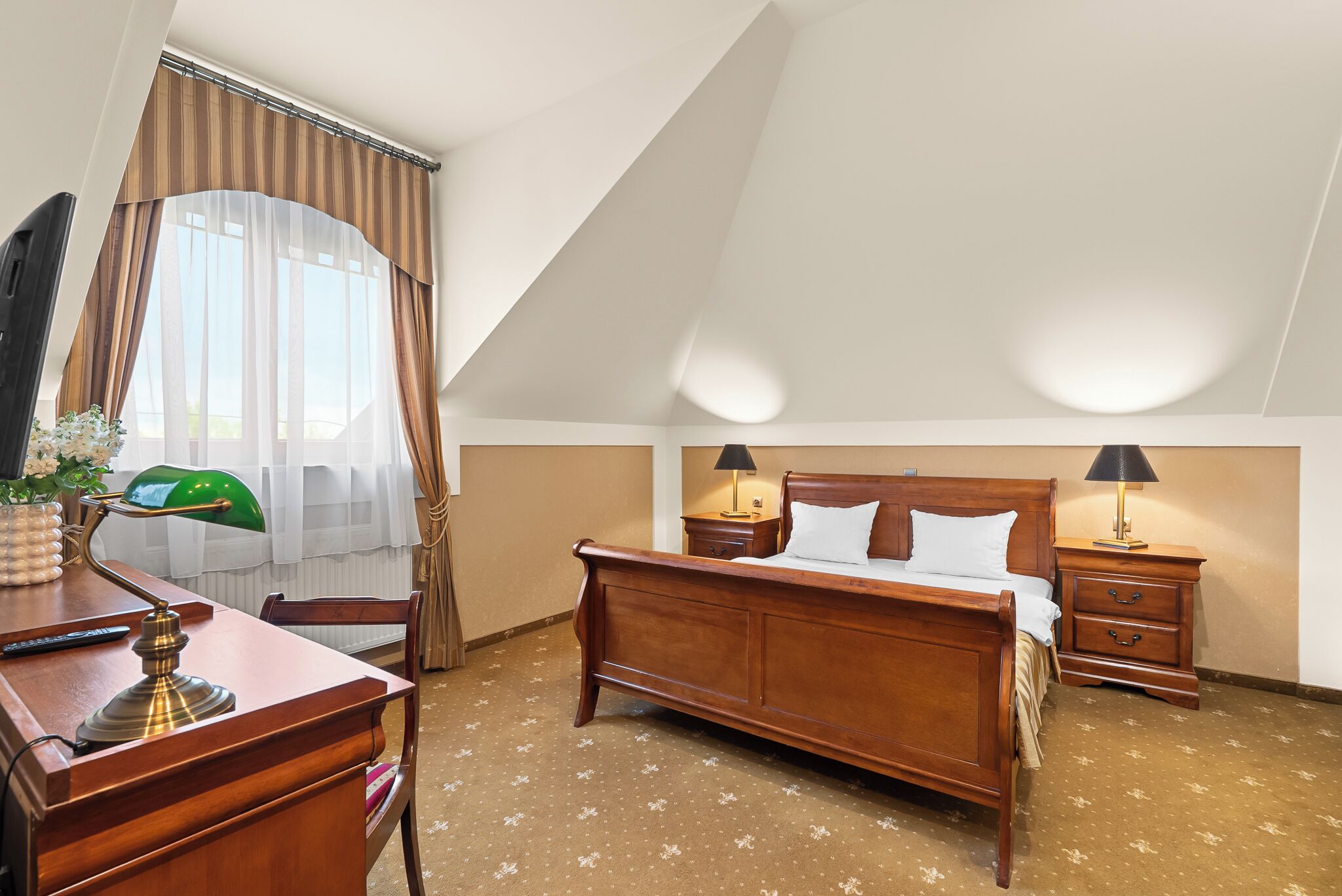 A hotel room with a wooden bed, two bedside tables with lamps, a desk with a lamp, a chair, and a window with curtains.