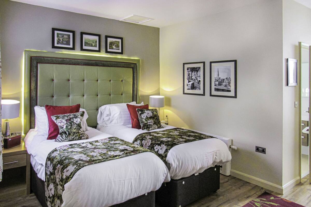 A hotel room with two beds, a green upholstered headboard, bedside lamps, and framed pictures on the wall.
