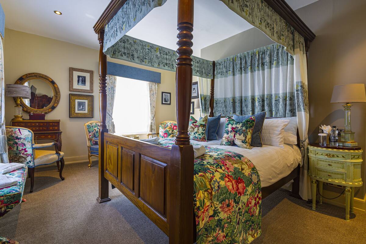 A four-poster canopy bed with floral bedding, a wooden nightstand with a lamp, and a chair with a floral cushion.