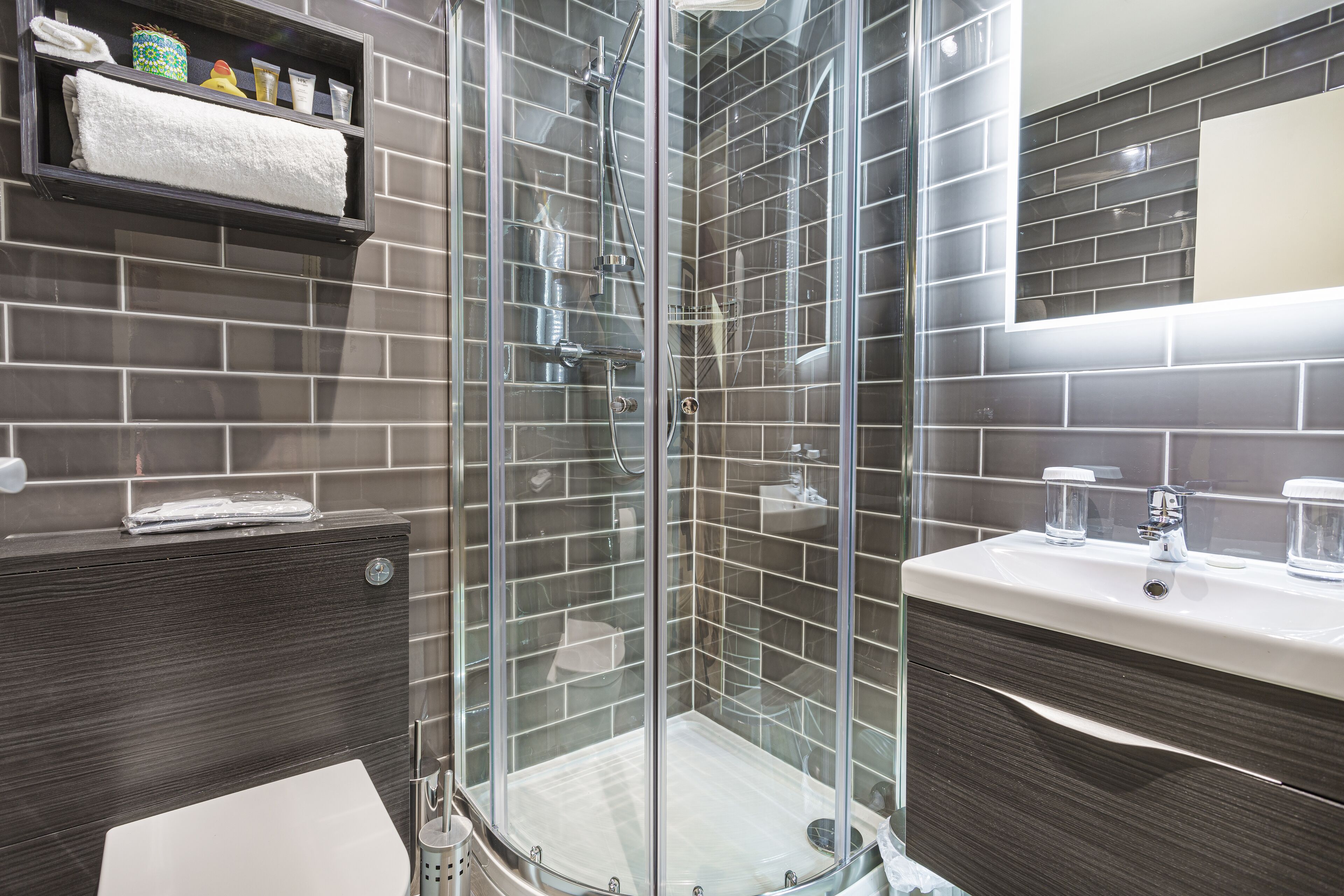 Une salle de bain moderne avec une douche à parois de verre, des murs recouverts de carreaux foncés et un lavabo blanc.