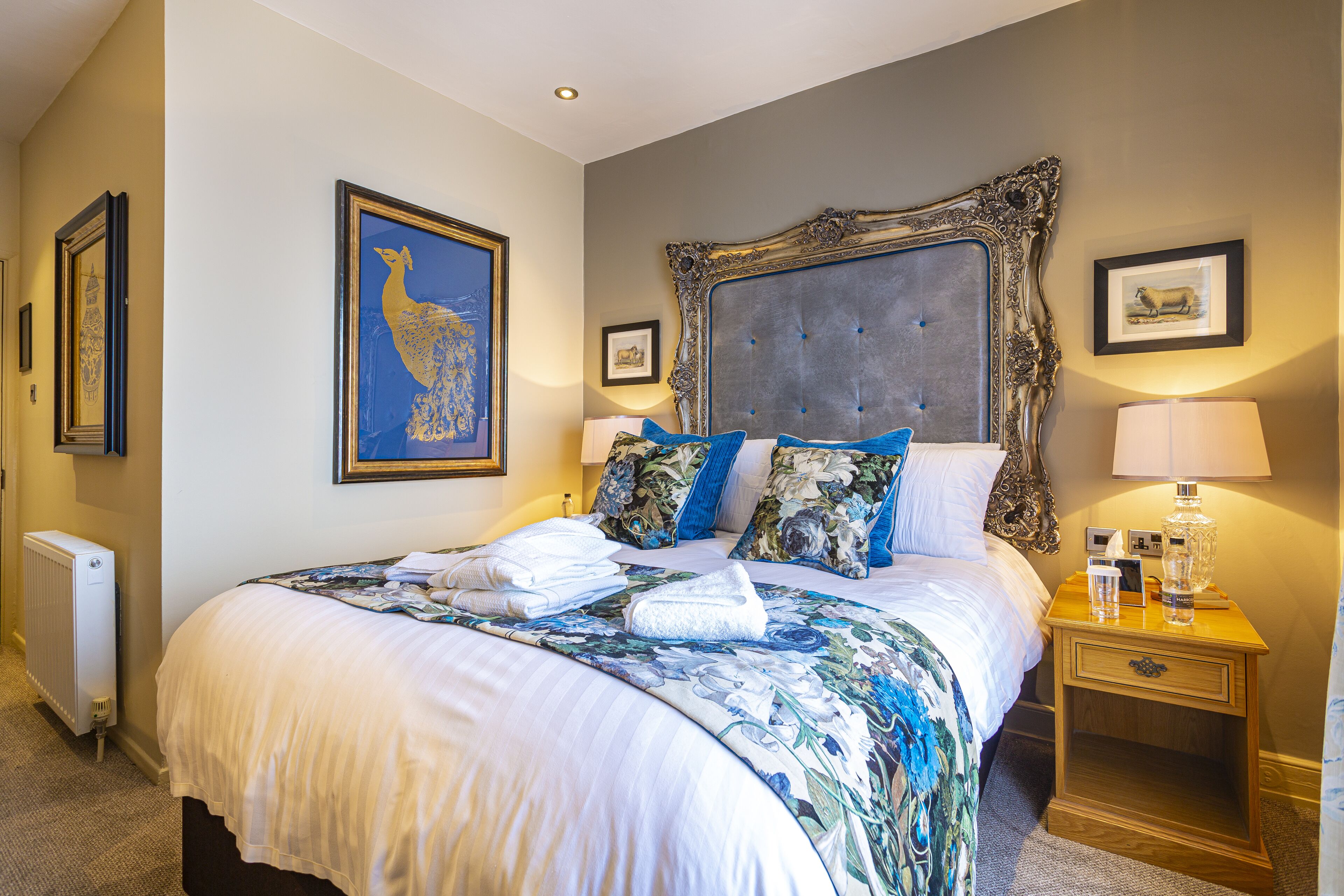 A neatly made bed with a floral bedspread, a large ornate headboard, and a bedside table with a lamp and a bottle.
