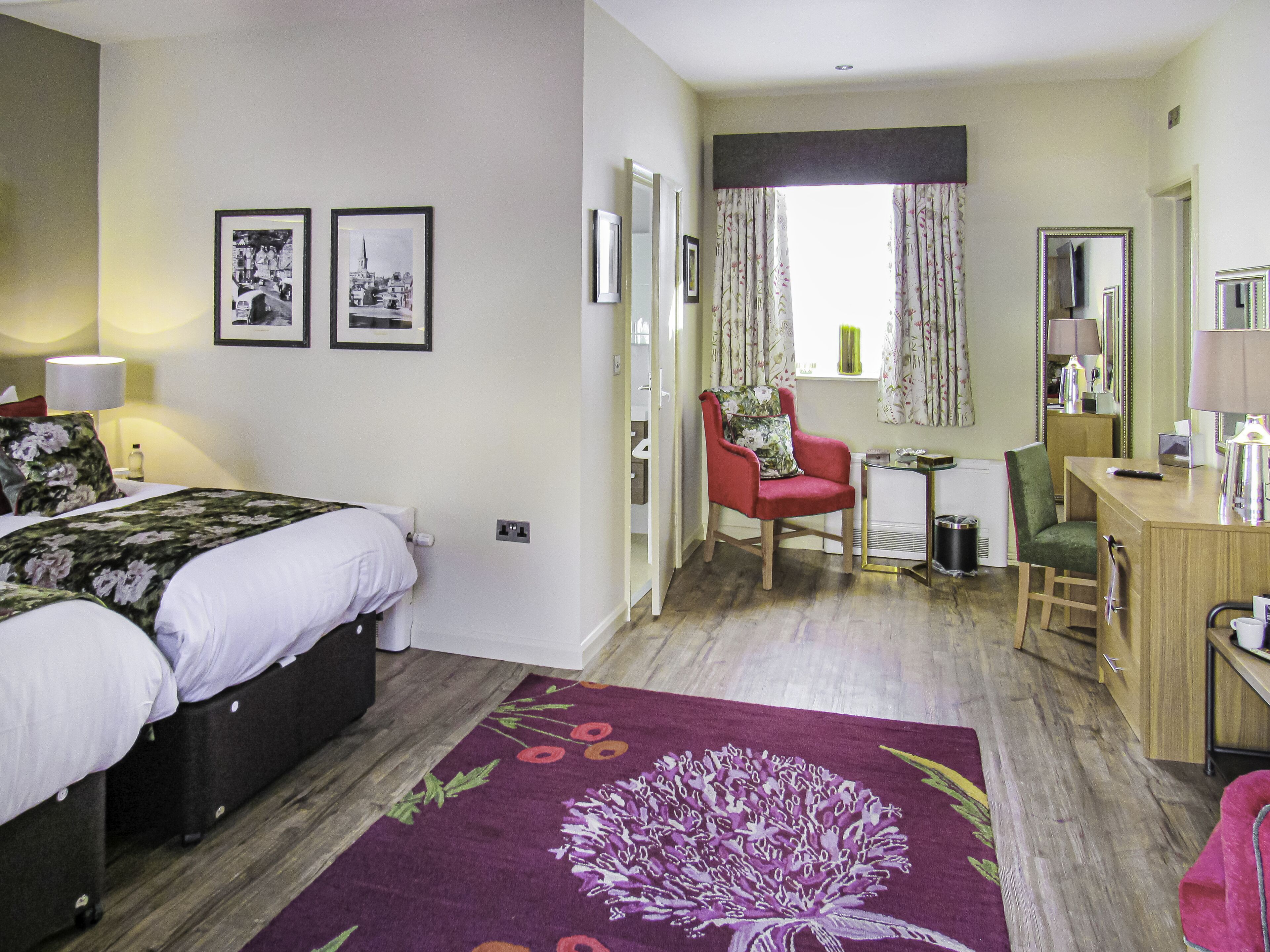 A hotel room with a bed, a desk, a chair, a mirror, and two framed pictures on the wall.