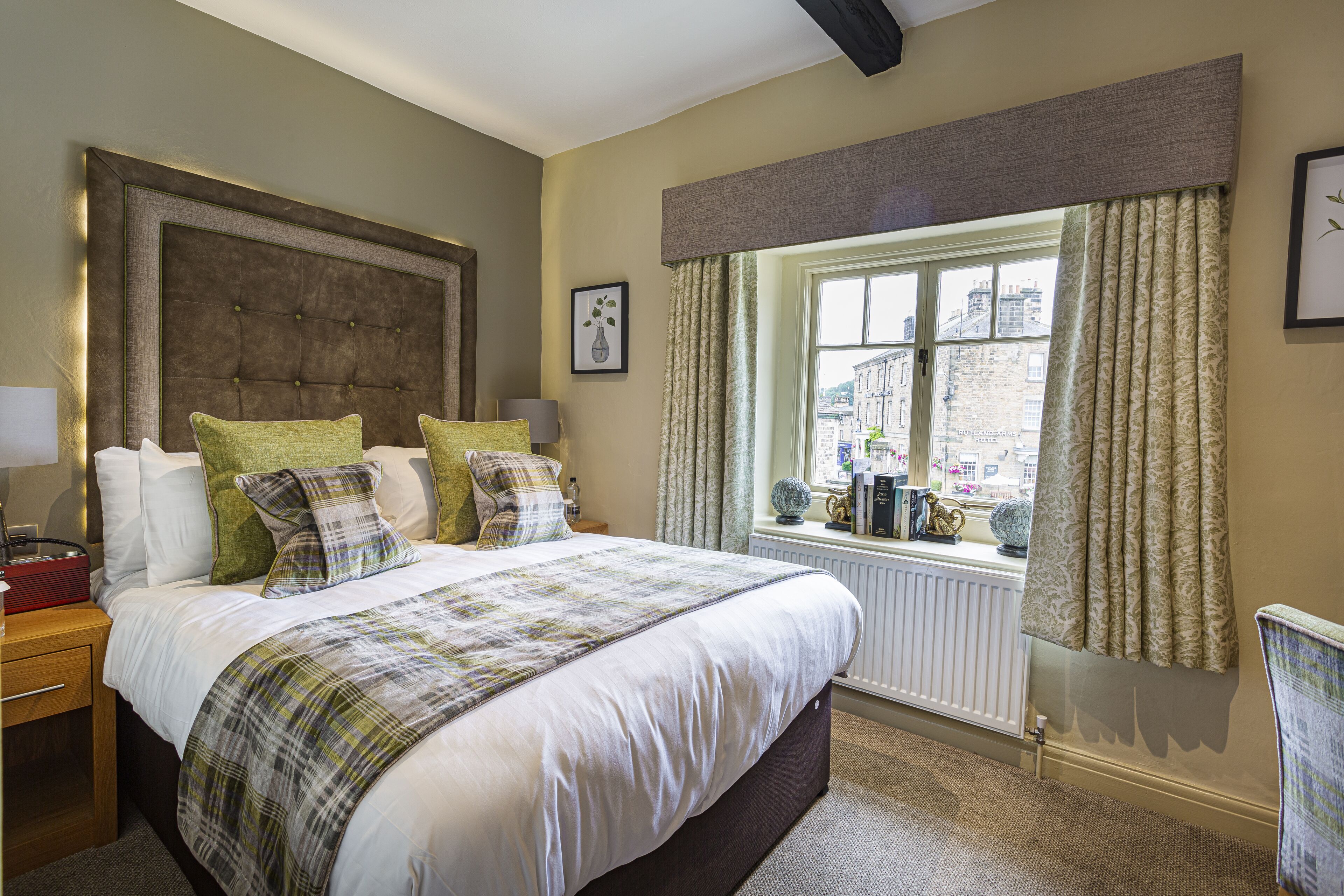 A neatly made bed with a plaid bedspread, a large headboard, and multiple pillows. There is a window with curtains, a radiator, and a view of buildings outside.
